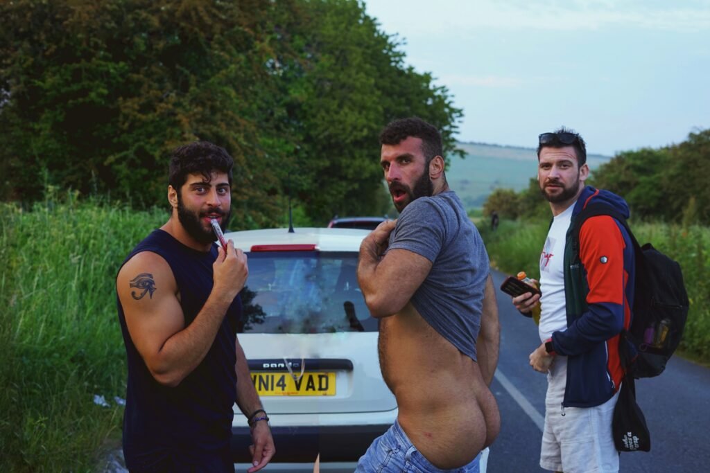 Three men pose next to a car, one of them standing naked in front, creating an unexpected and amusing visual.