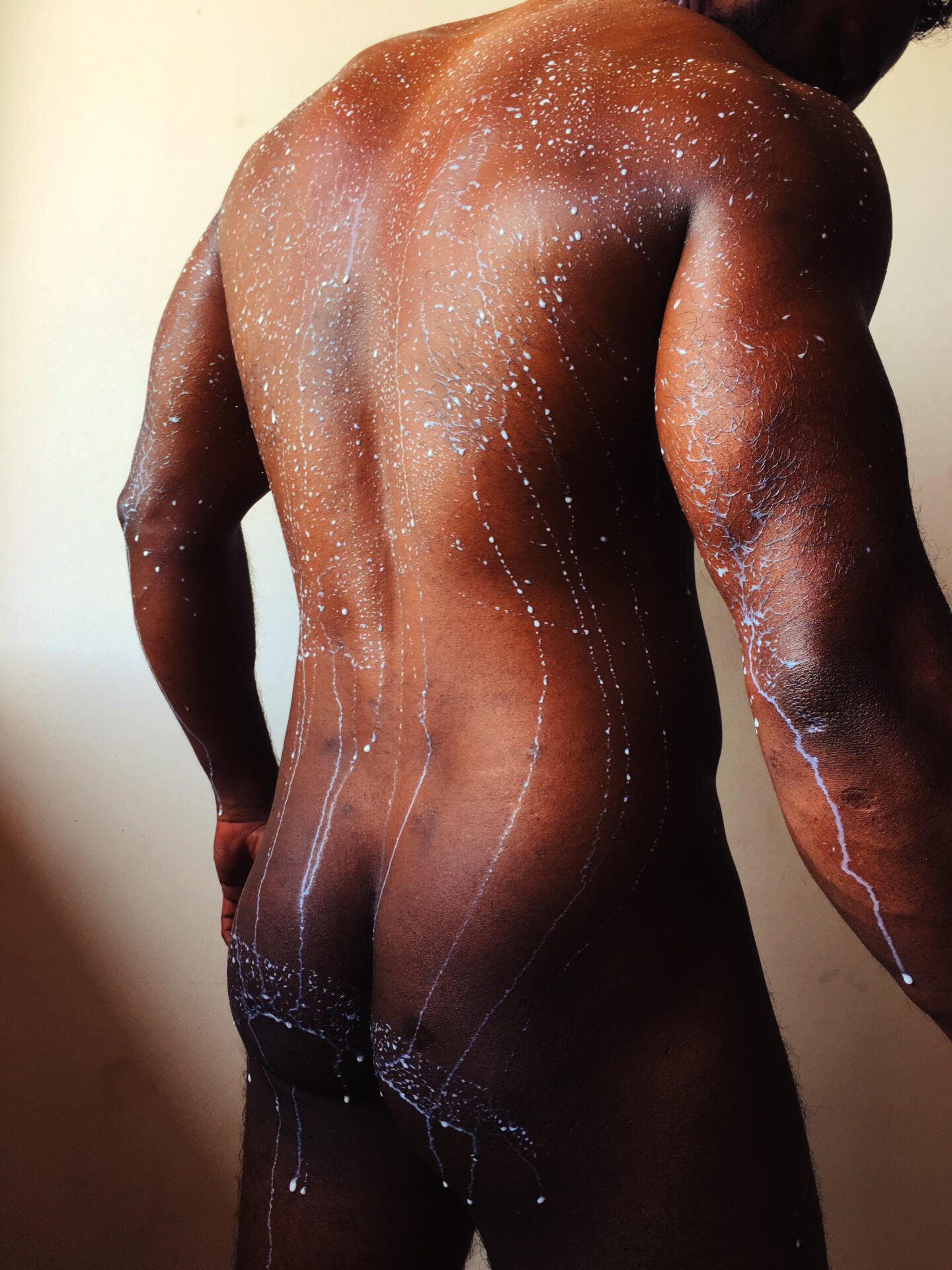 A nude man poses as milk droplets cascade down his skin, creating a striking visual effect.