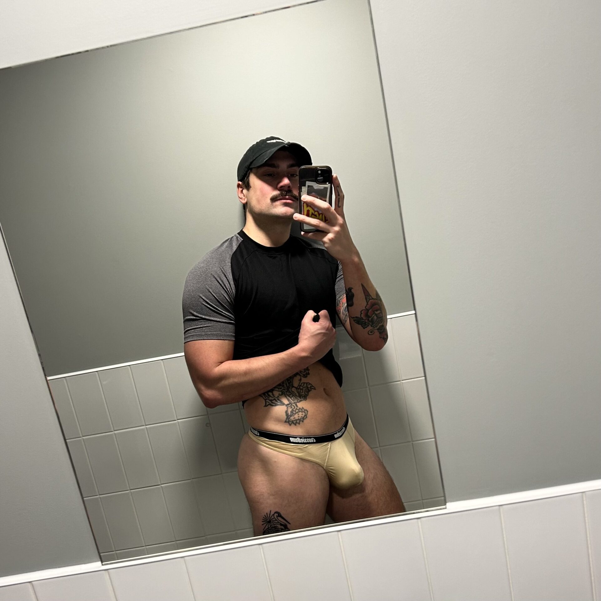 A guy in a thong snaps a selfie in the bathroom mirror, showing off his fun side with a confident grin.